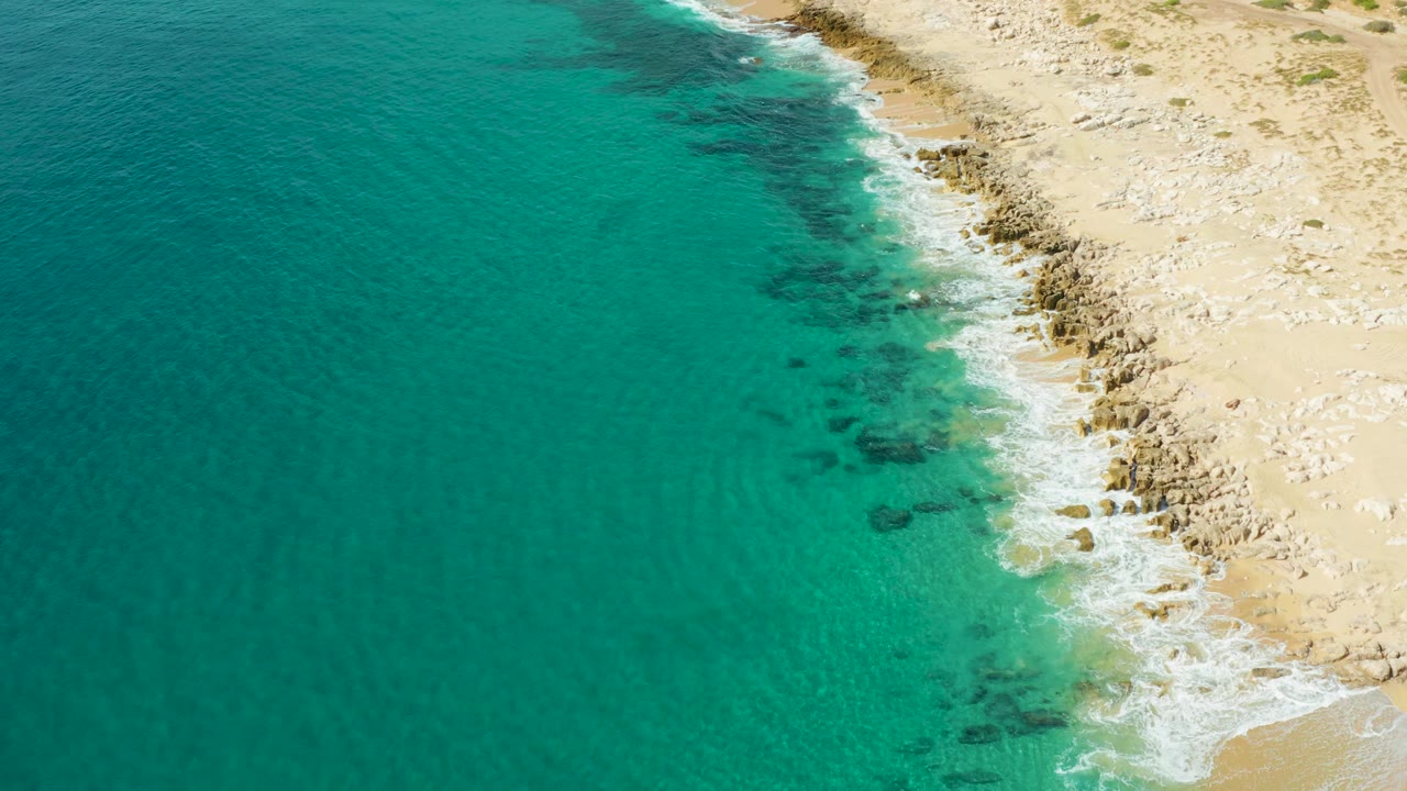 Aerial frame of clear turquoise water, dark underwater rock patches, and white foam along the Laguna Beach Los Cabos shore, with no visible people.