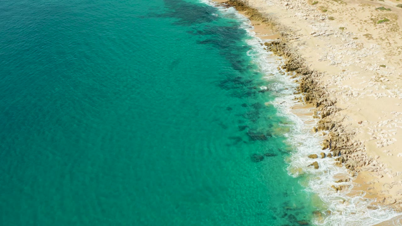 Turquoise sea fills the left side while a pale rocky beach edge runs down the right at Laguna Beach Los Cabos, with no visible people.