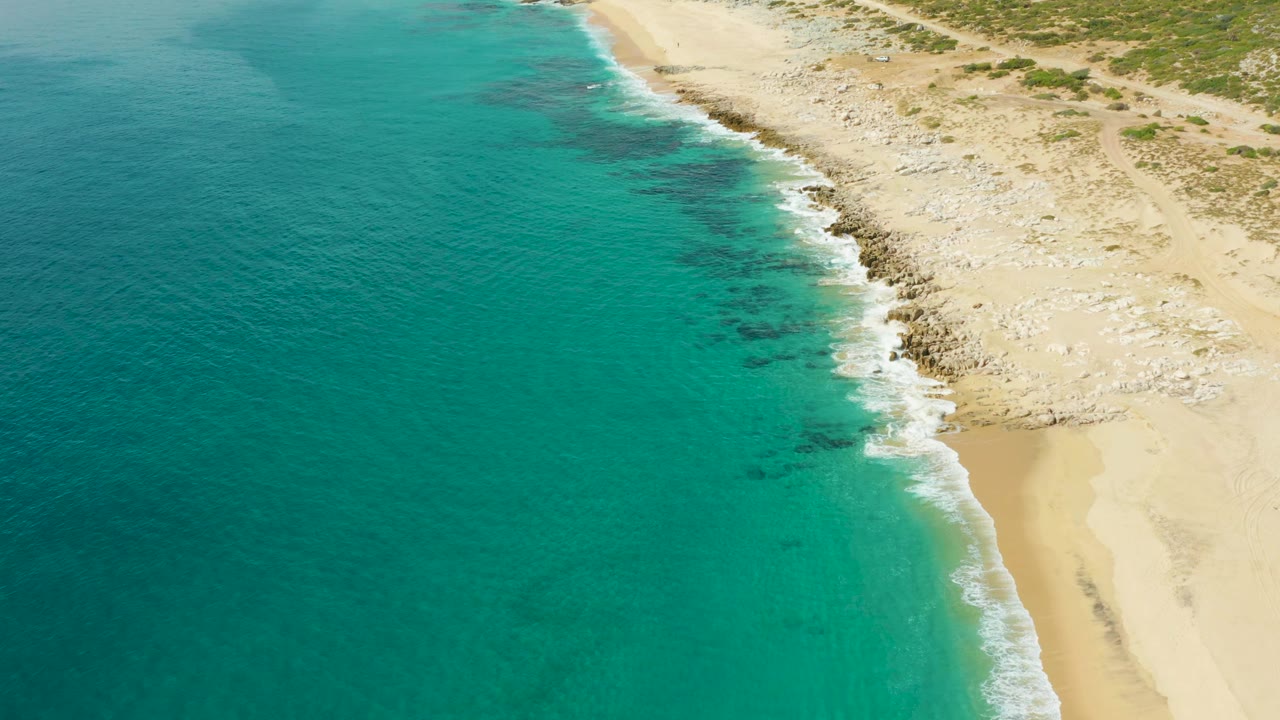 Aerial frame from Laguna Beach Los Cabos showing white foam building along the rocky edge while shallow turquoise water spreads beside the sand, with no visible people.