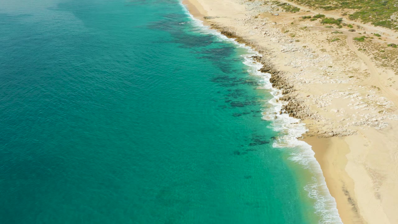 Laguna Beach Los Cabos shoreline seen from above, with a broader patch of clear teal water beside a curved sandy beach and rocky surf line, with no visible people.