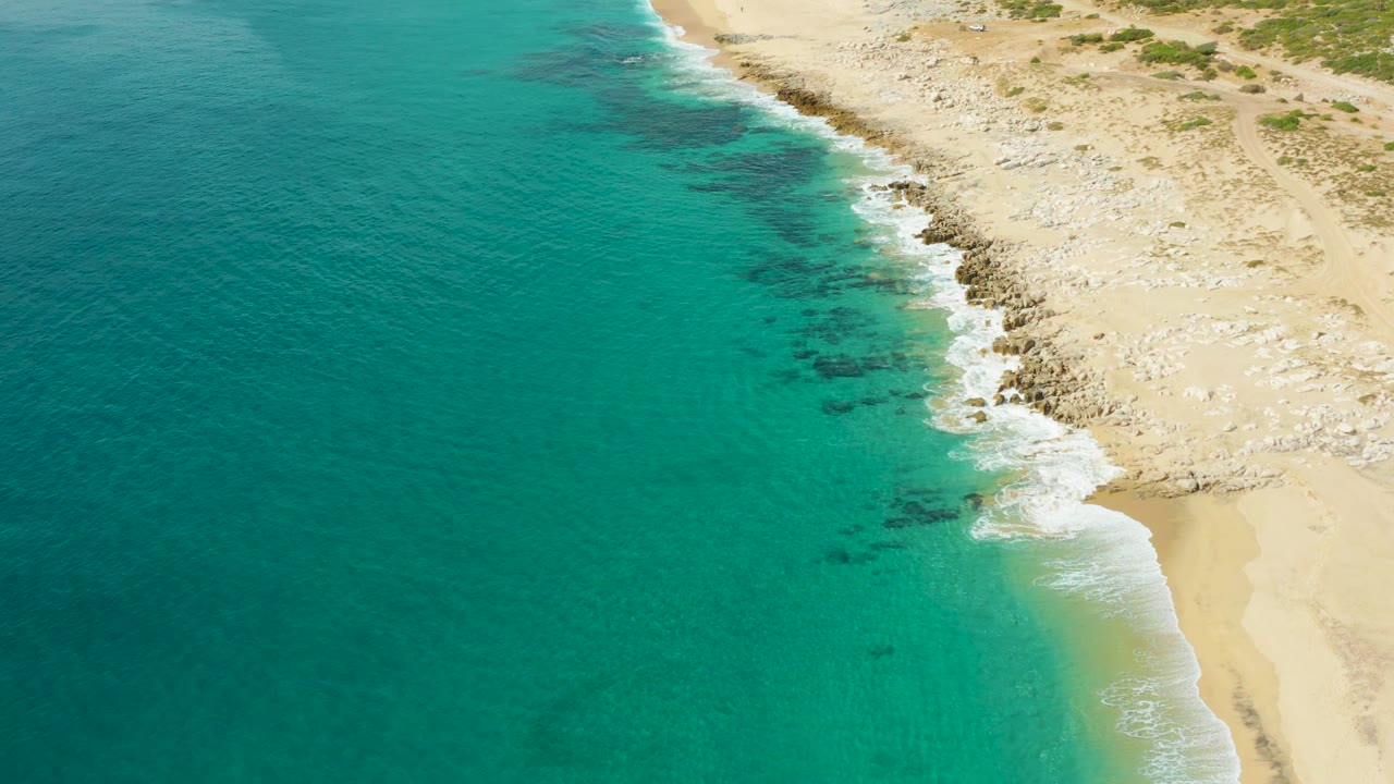 Aerial view at Laguna Beach Los Cabos with wide turquoise ocean filling most of the frame and white foam tracing the rocky shoreline near pale sand, with no visible people.
