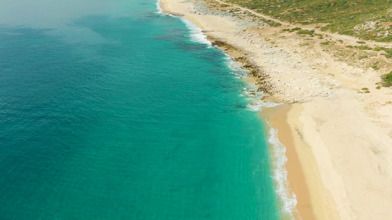 Aerial frame over Laguna Beach Los Cabos with clear blue-green water, a narrow band of white surf, pale sand, exposed rocks, scrubland, and no visible people.