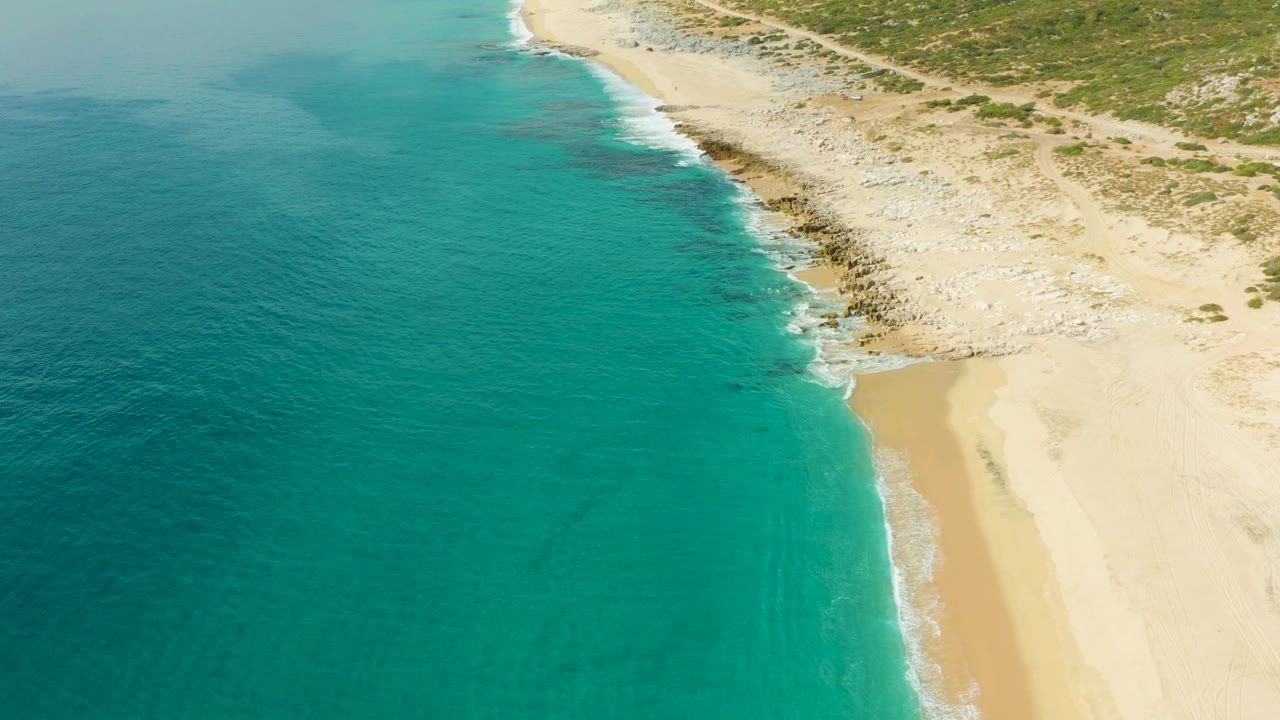 Mid-clip aerial view of Laguna Beach Los Cabos showing a broad turquoise water area, foamy waves along the sand, light rocks above the waterline, and no visible people.