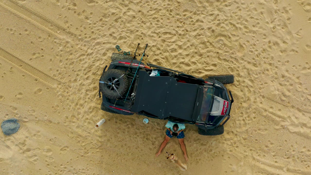 Overhead frame from Laguna Beach Los Cabos shows a black off-road vehicle on tan sand with a person standing near its side and gear visible on the rear rack.