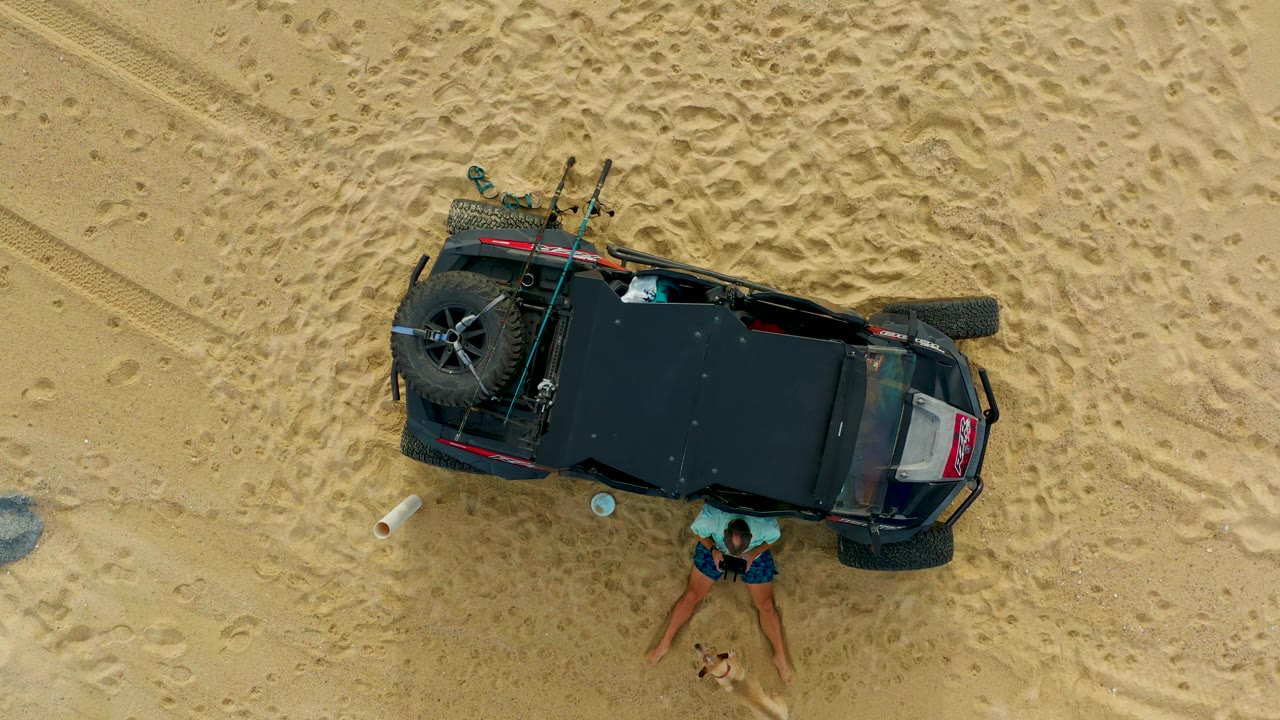 In Laguna Beach Los Cabos, the overhead frame shows the off-road vehicle angled across rippled sand, with the person below the side door area and loose items on the ground nearby.