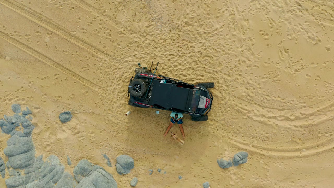At 0:00 in Laguna Beach Los Cabos, Baja California Sur, a black 4x4 is seen from overhead on beige sand with one person standing beside the vehicle near pale rocks.