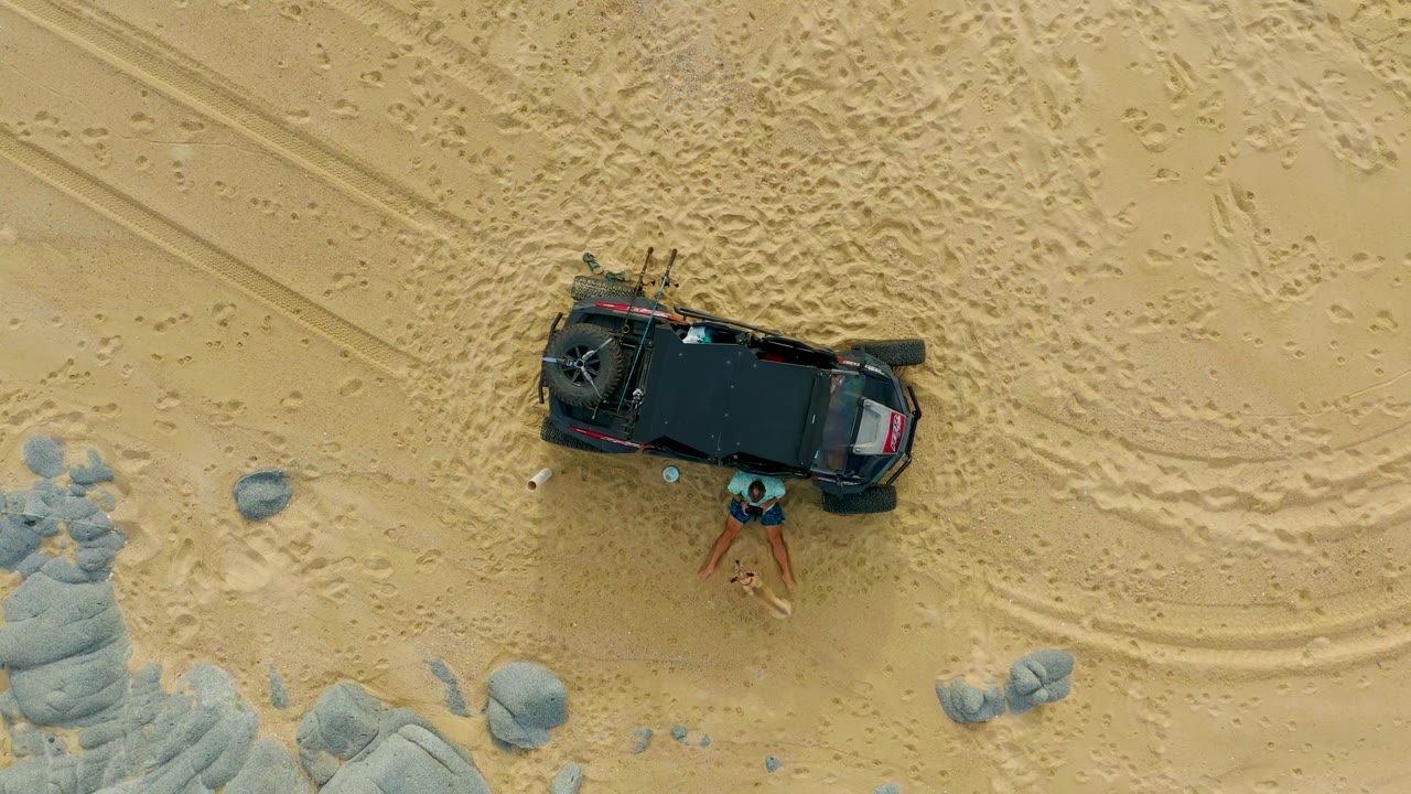 At 0:02 in Laguna Beach Los Cabos, the black off-road vehicle remains centered on sandy terrain while the visible person stands just below the vehicle.