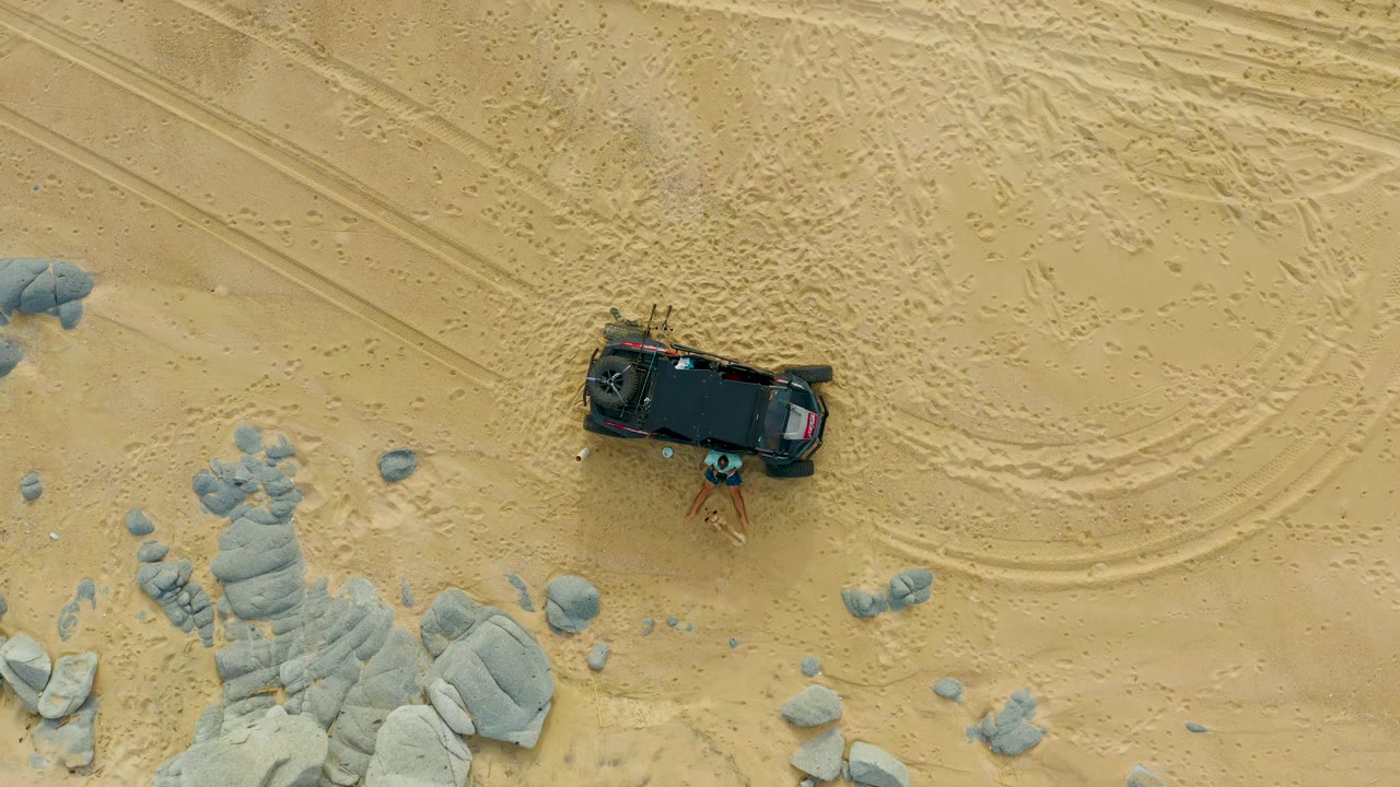 Overhead frame from Laguna Beach Los Cabos showing a black SUV parked on pale sand beside gray rock clusters, curved tire tracks, and small visible people near the vehicle.