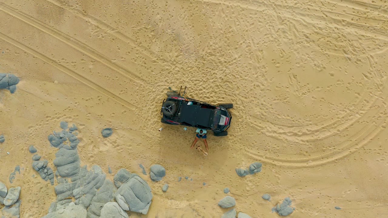 The 0:02 frame shows the black SUV in Laguna Beach Los Cabos beside scattered gray rocks, with faint footprints or tire marks in the sand and people visible near the vehicle.