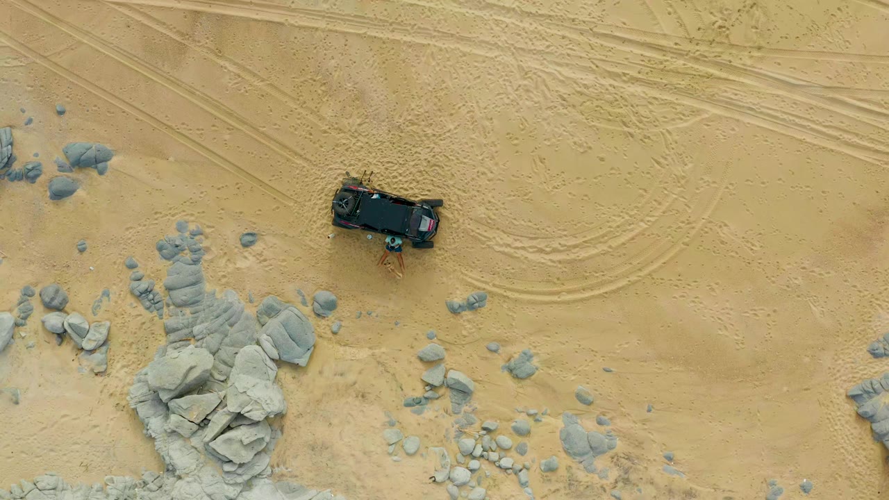 Overhead frame from Laguna Beach Los Cabos showing a dark truck on beige sand above scattered gray rocks and tire tracks, with no visible people.