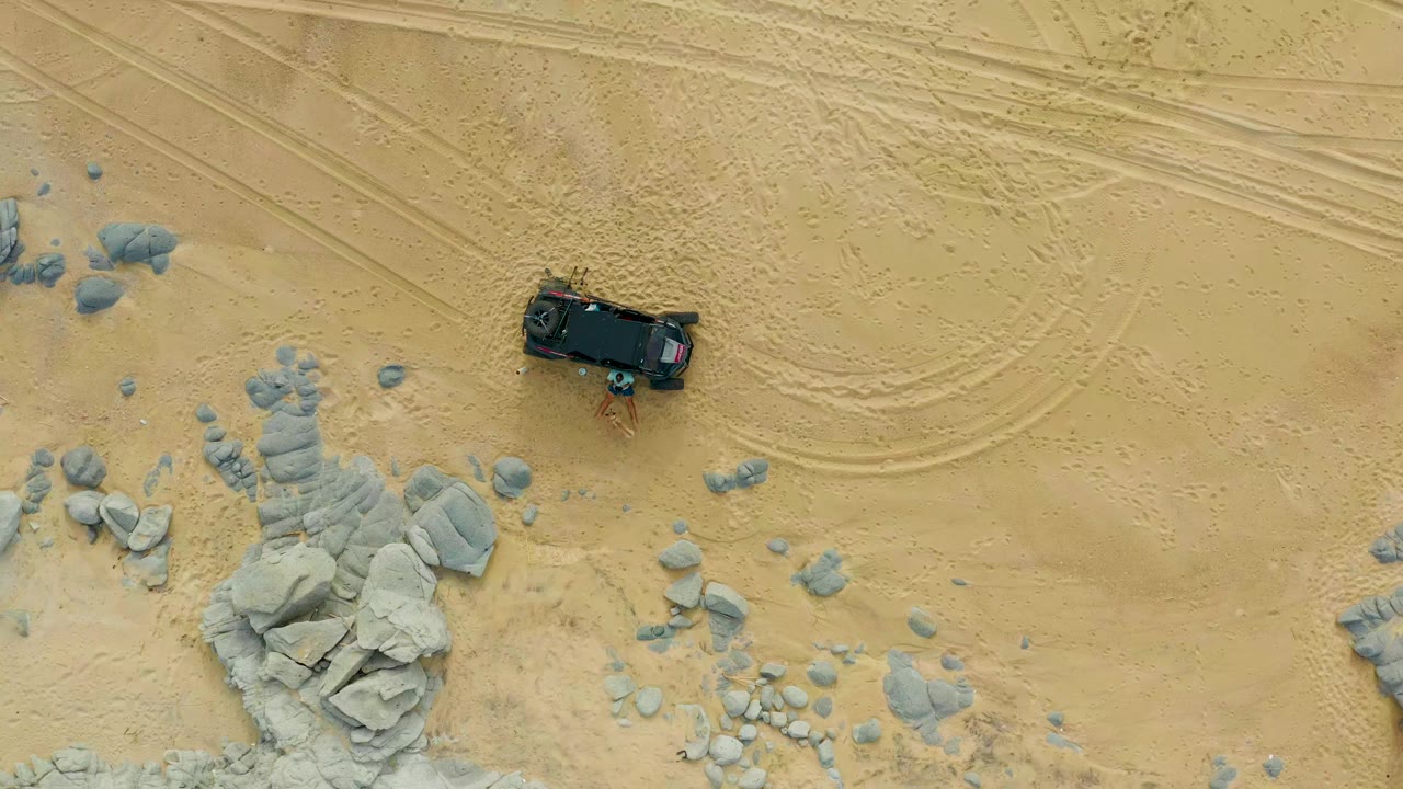 Aerial view in Laguna Beach Los Cabos with the dark truck slightly farther across beige sand, gray rock clusters below, and no visible people.