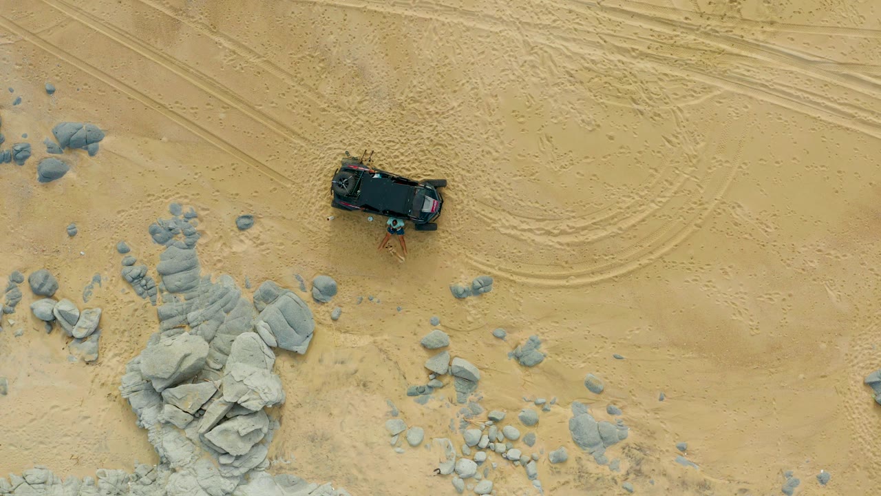 Overhead Laguna Beach Los Cabos frame showing the truck near the upper right, curved tire tracks across the sand, scattered gray rocks, and no visible people.