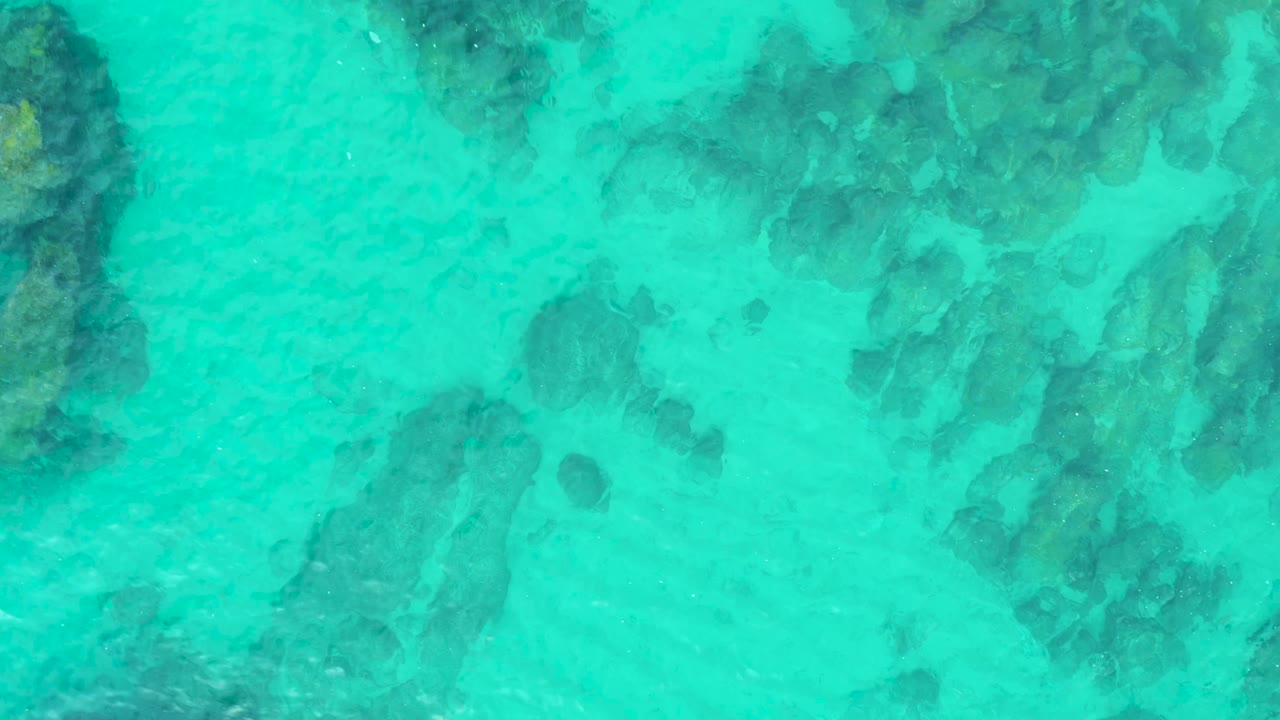 Clear blue-green water reveals scattered underwater rocks and pale sand at Laguna Beach Los Cabos, with ripples softening the rock edges and no visible people.