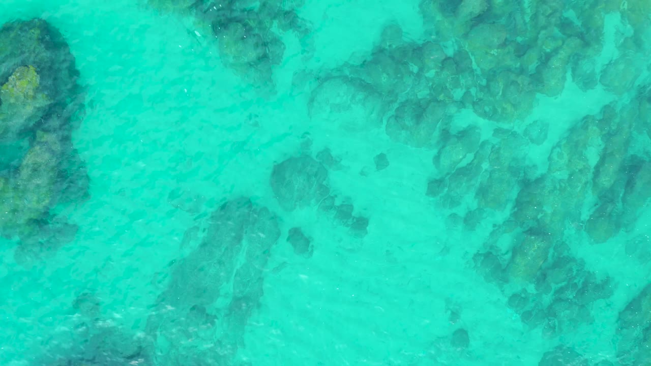 Turquoise ocean surface covers darker rock patches beneath the water at Laguna Beach Los Cabos, with a larger rock area toward the upper right and no visible people.