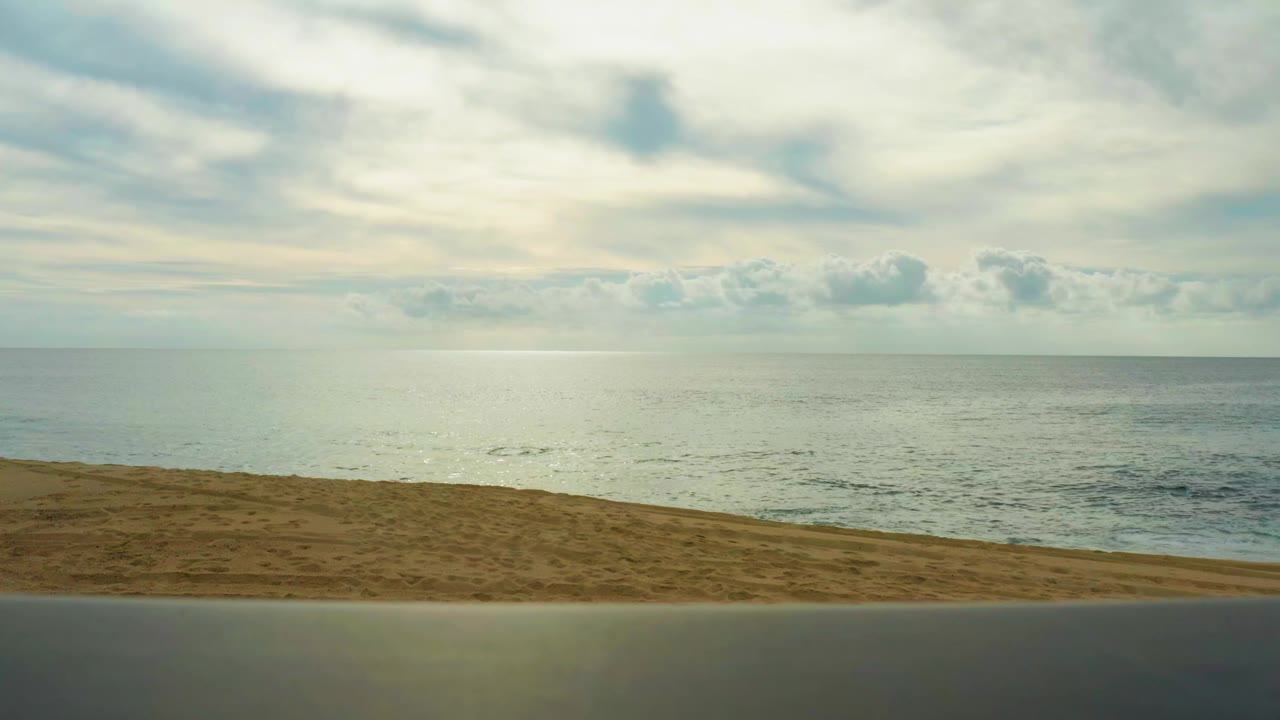Laguna Beach Los Cabos shoreline one second in, with the foreground road below a sloped sand strip and pale ocean under a bright cloudy sky, no visible people.