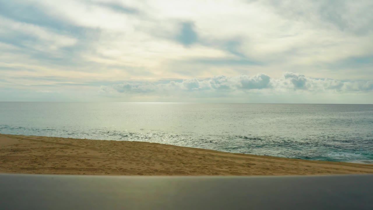 Mid-clip frame from Laguna Beach Los Cabos showing the road band, sandy beach, faint incoming waves, distant horizon, soft cloud cover, and no visible people.