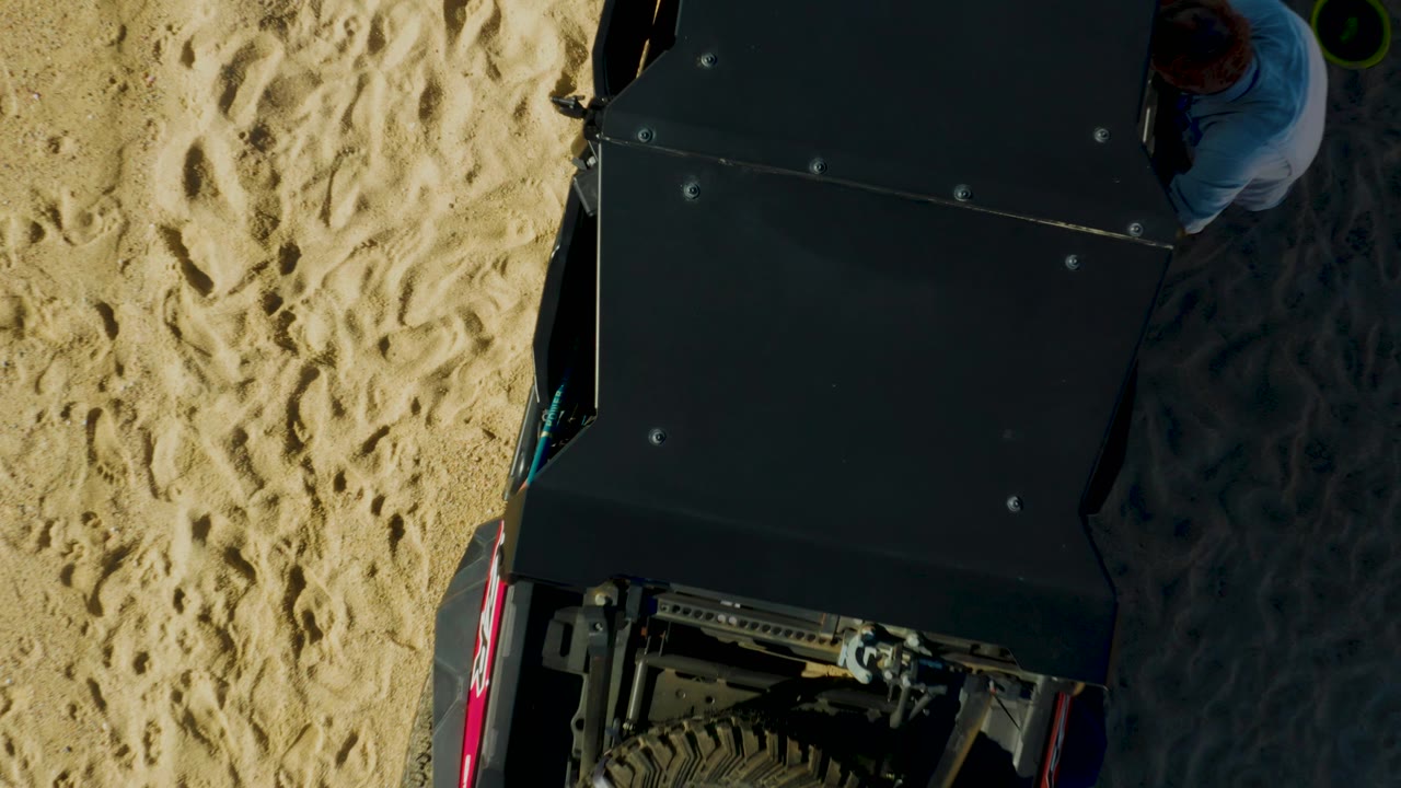 Overhead frame in Laguna Beach Los Cabos showing a dark off-road vehicle or buggy on pale sand, with a person partly visible beside the upper right edge.