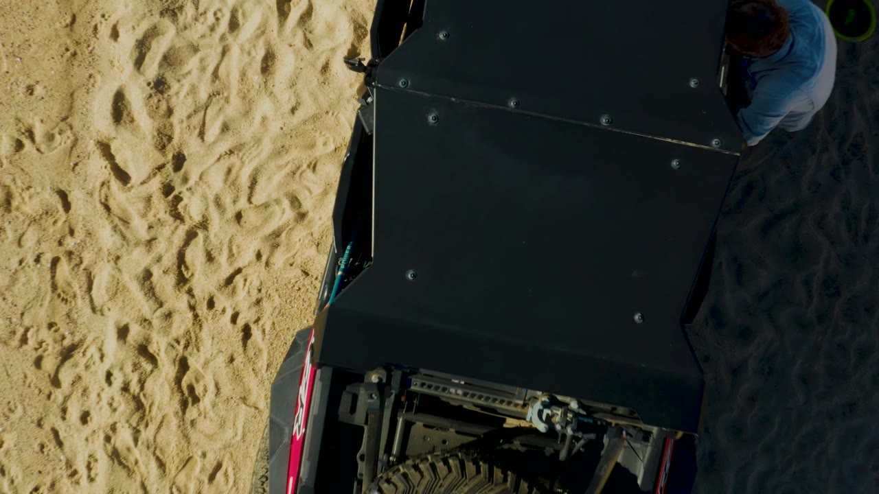 Overhead view of the same dark vehicle on sand with part of a person at the edge.