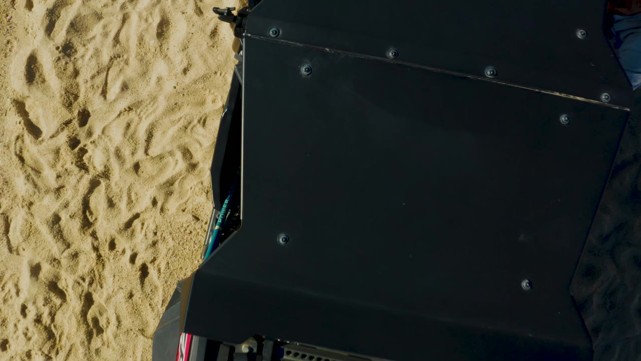 Close overhead frame in Laguna Beach Los Cabos showing the dark vehicle roof filling most of the image over textured sand, with no visible people in this frame.