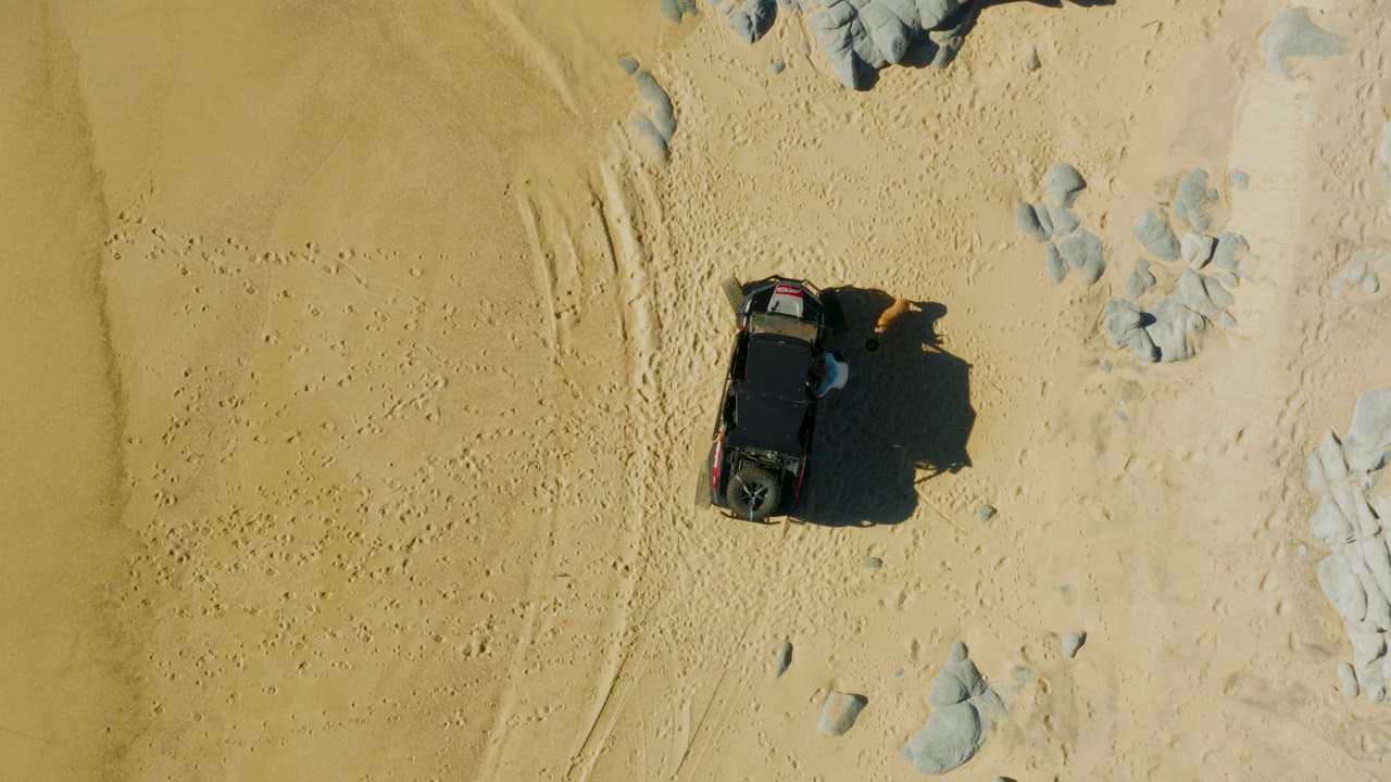 Overhead frame from Laguna Beach Los Cabos, Baja California Sur showing a dark SUV on pale sand with tire marks, scattered rocks, and no visible people.