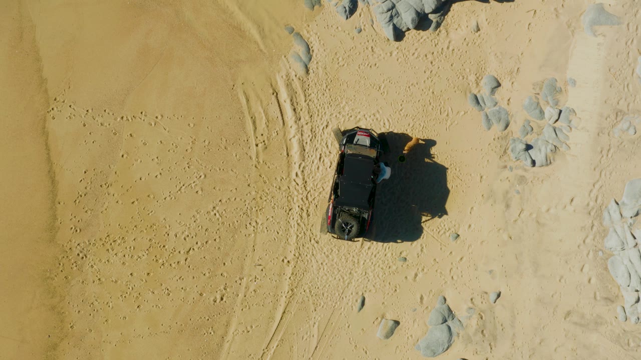 Dark SUV viewed from above on sandy ground beside tire tracks and light rocks.