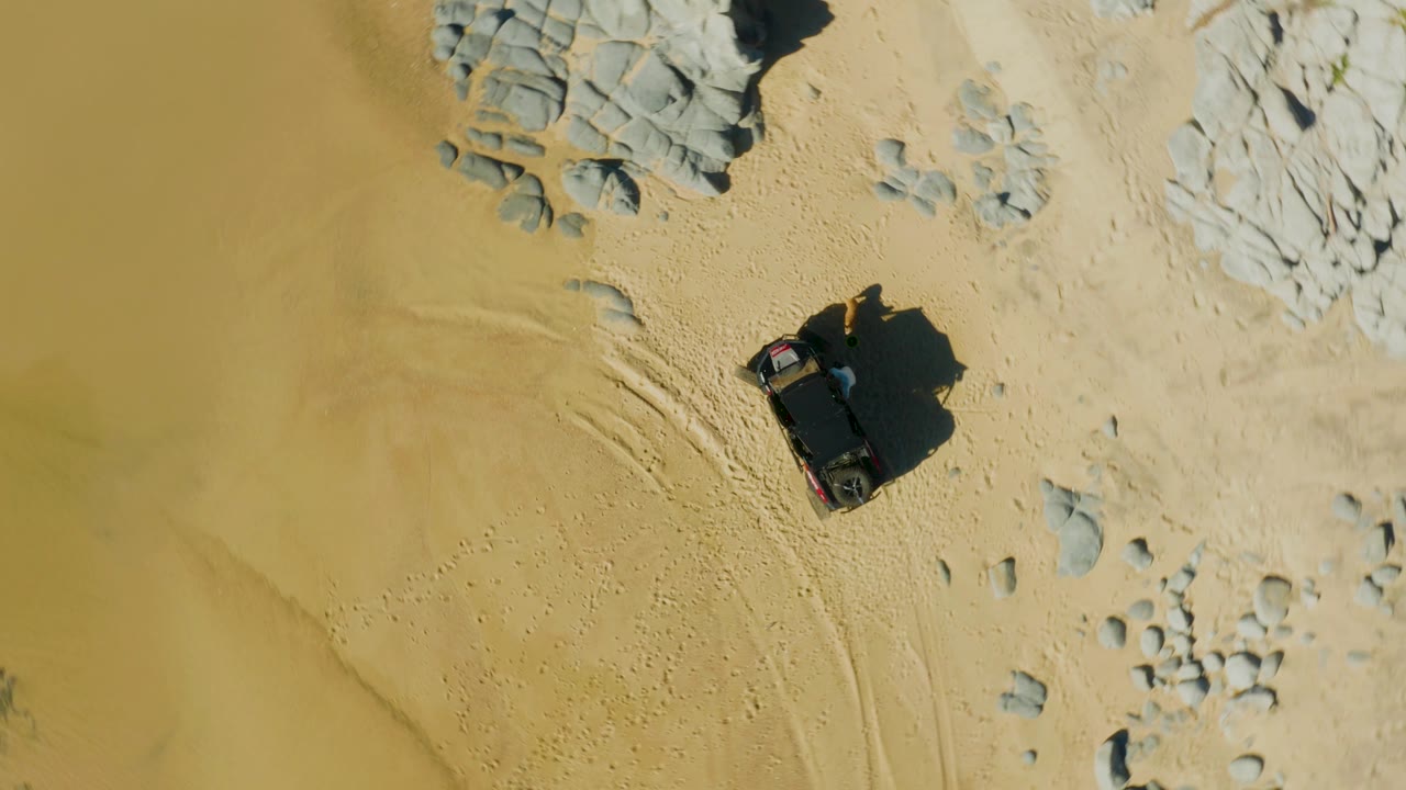 Top-down drone view of the SUV near pale rock outcrops and smoother yellow sand, with its shadow falling to one side and no visible people.