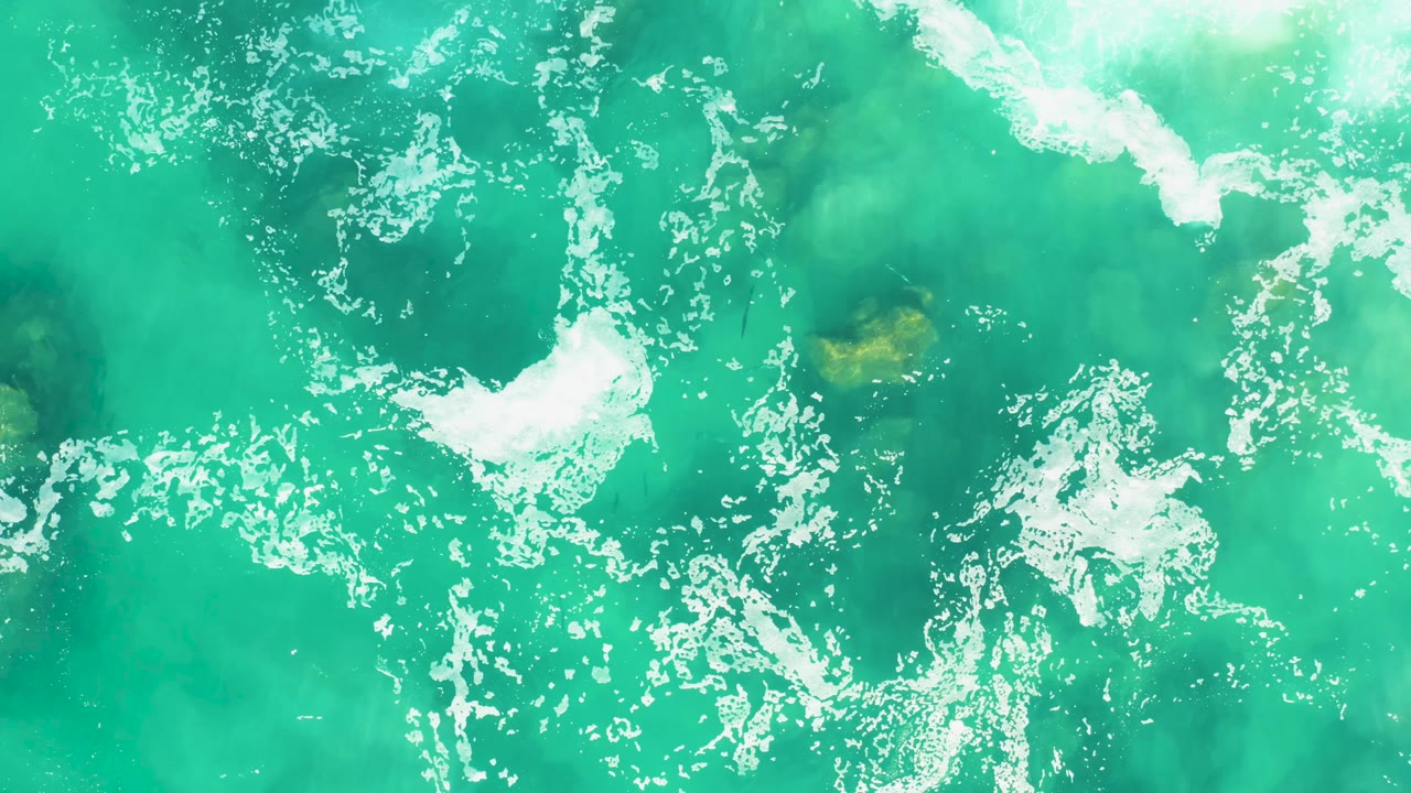 Aerial view over Laguna Beach Los Cabos water where a bright white foam streak crosses turquoise sea above faint seabed color, with no visible people.
