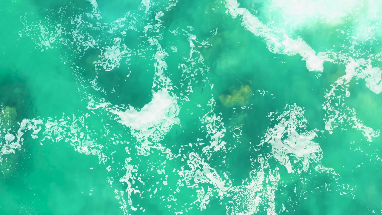 Top-down Laguna Beach Los Cabos ocean frame with white foam clusters spread across green-blue water and a small darker patch beneath the surface, with no visible people.