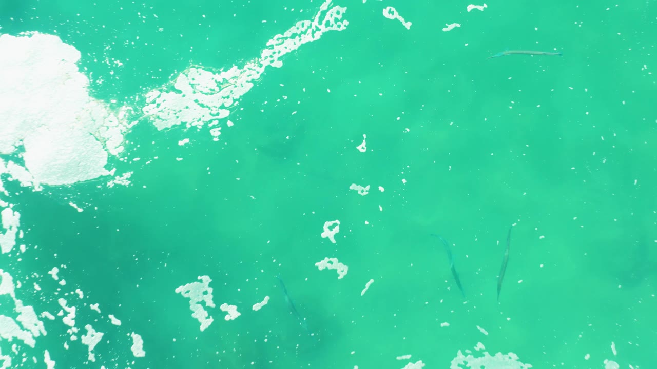 A larger white foam patch enters the upper left of the Laguna Beach Los Cabos water view, with faint fish visible under the turquoise surface and no visible people.