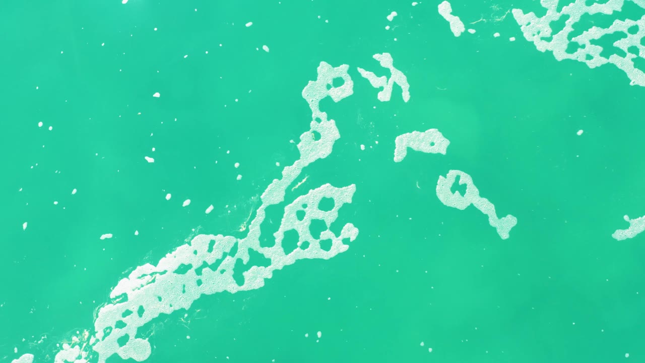 At Laguna Beach Los Cabos, a larger irregular white foam pattern spreads across the lower left of the turquoise ocean surface, with no visible people.