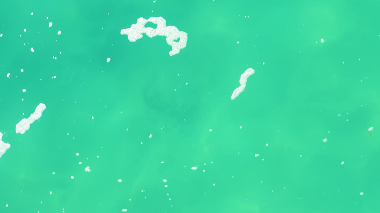 Overhead frame from Laguna Beach Los Cabos with larger white foam clusters on bright green-blue water and faint darker patches beneath the surface, no visible people.