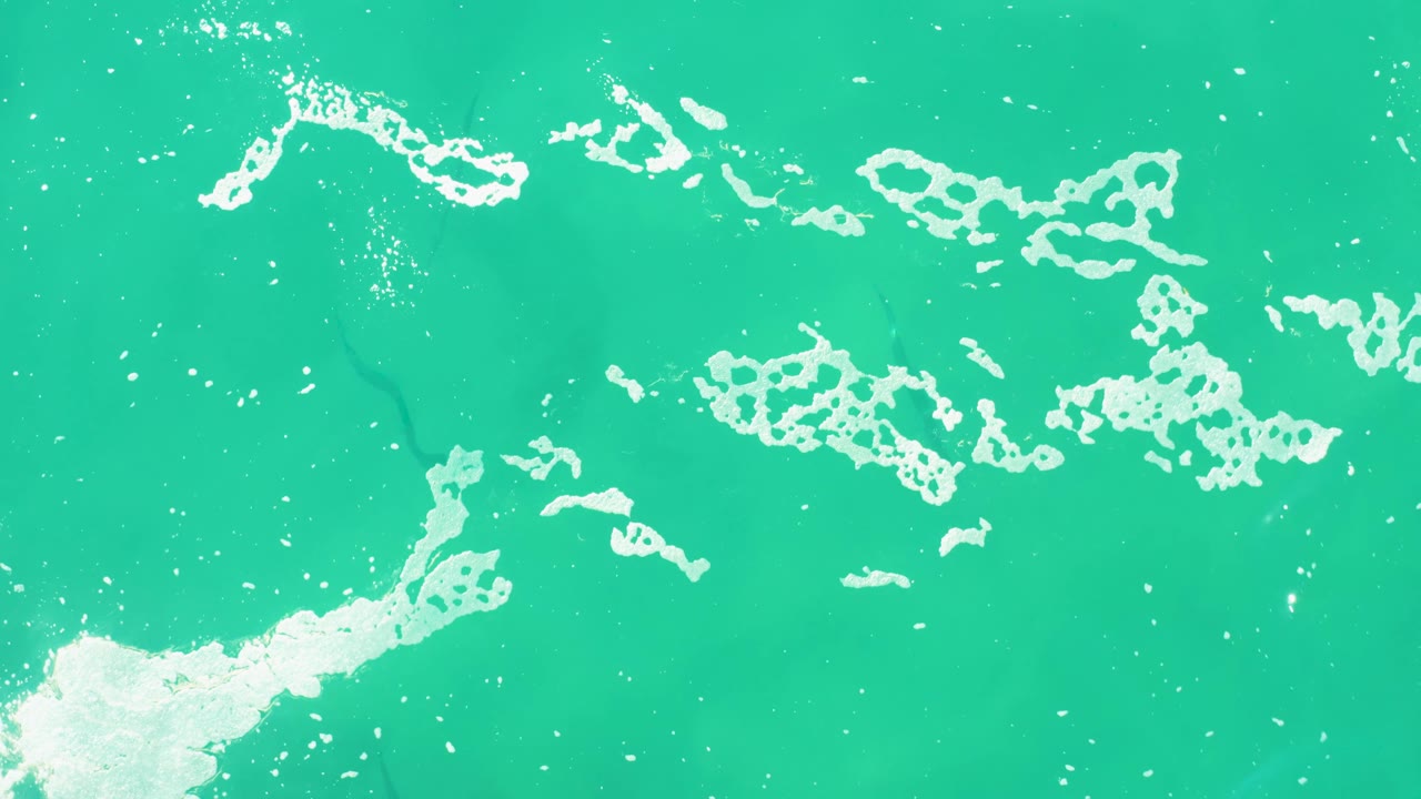 White surf foam forms broken bands over turquoise water in an overhead Laguna Beach Los Cabos frame, with no visible people.