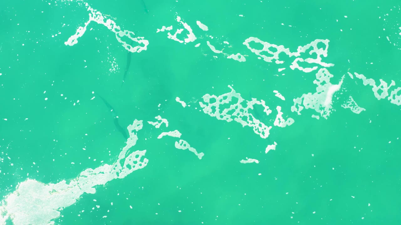 A larger white foam cluster stretches diagonally across turquoise water in the Laguna Beach Los Cabos aerial view, with no visible people.