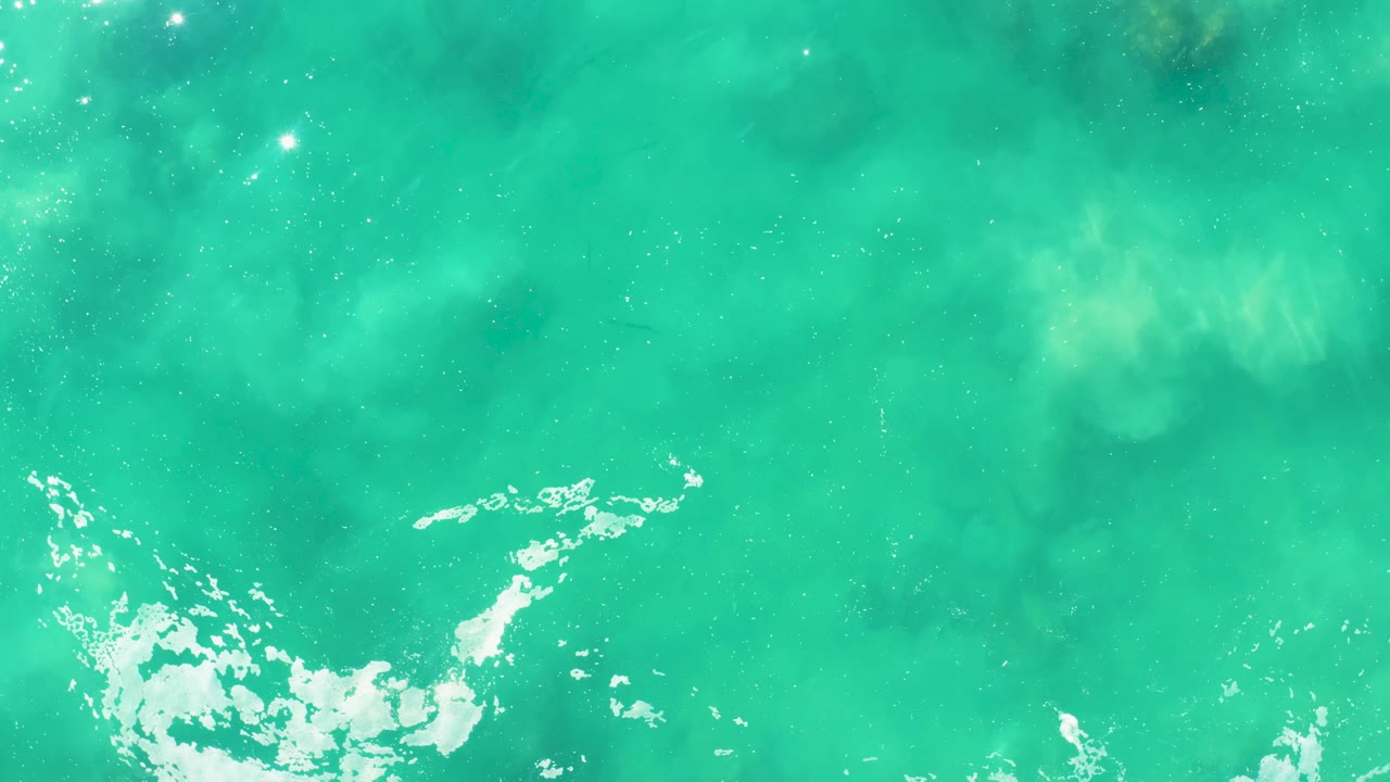 Overhead shallow water at Laguna Beach Los Cabos shows darker submerged shapes under turquoise water and a broken foam edge below, with no visible people.