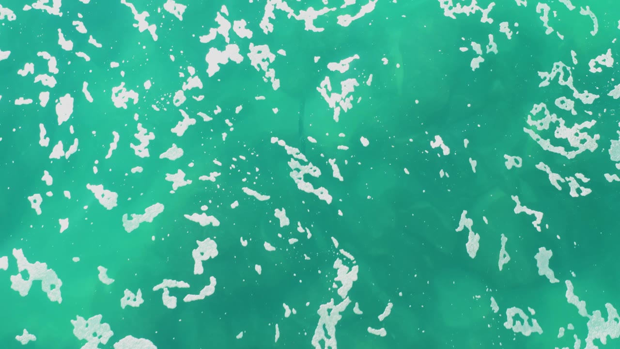 At one second, white foam flecks sit across bright turquoise shallow water in Laguna Beach Los Cabos, with no horizon, land, or visible people.