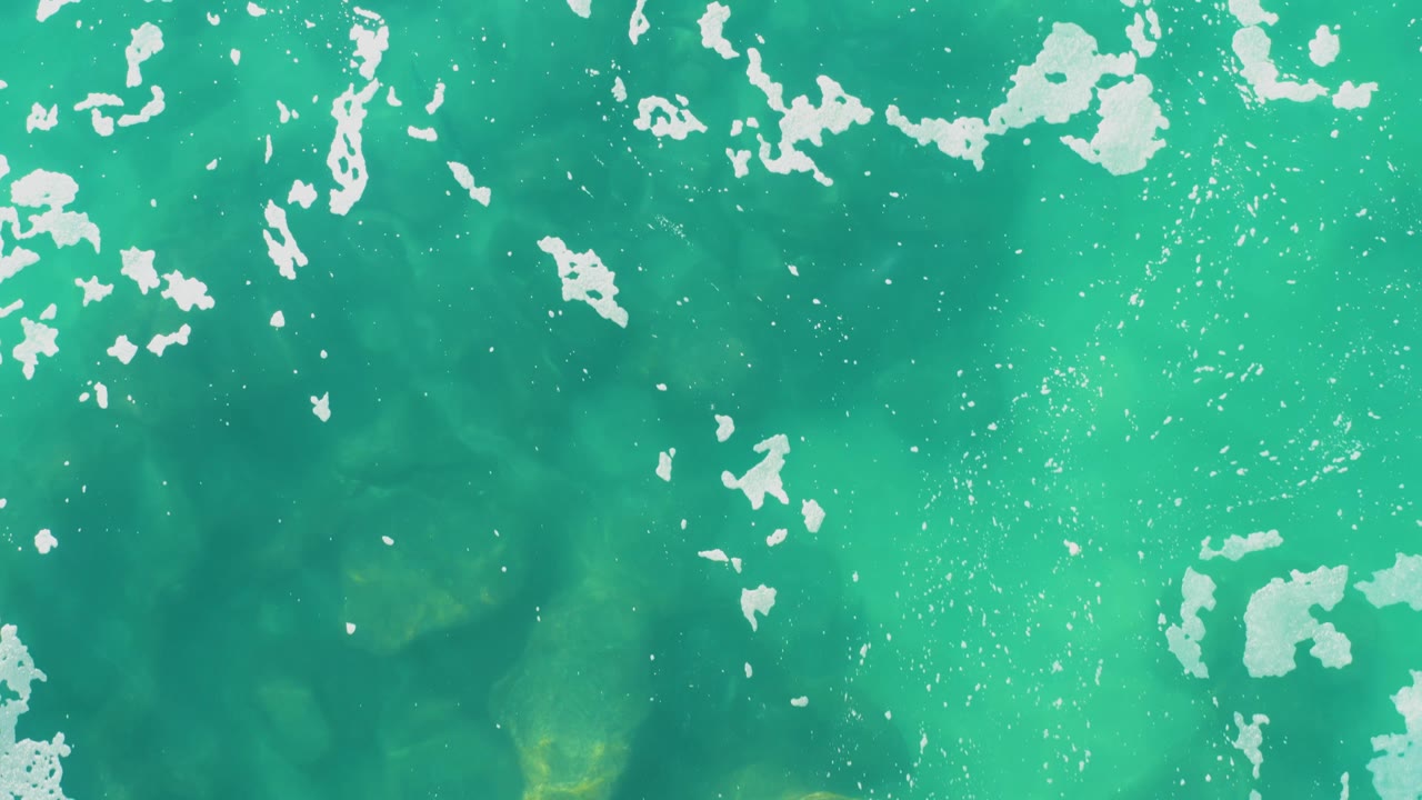 Overhead frame of clear turquoise water in Laguna Beach Los Cabos, with white foam bands crossing the upper edge and no visible people.