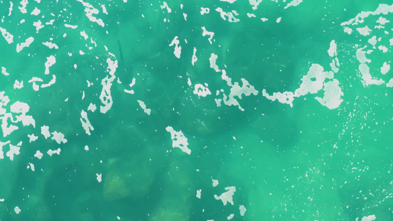 Overhead view of turquoise-green ocean at Laguna Beach Los Cabos, with scattered white foam across the upper half and no visible people.
