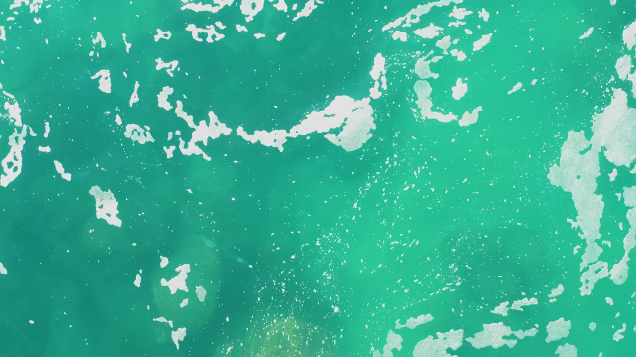 Overhead frame from Laguna Beach Los Cabos showing broken white foam bands on clear turquoise sea with faint darker underwater areas and no visible people.