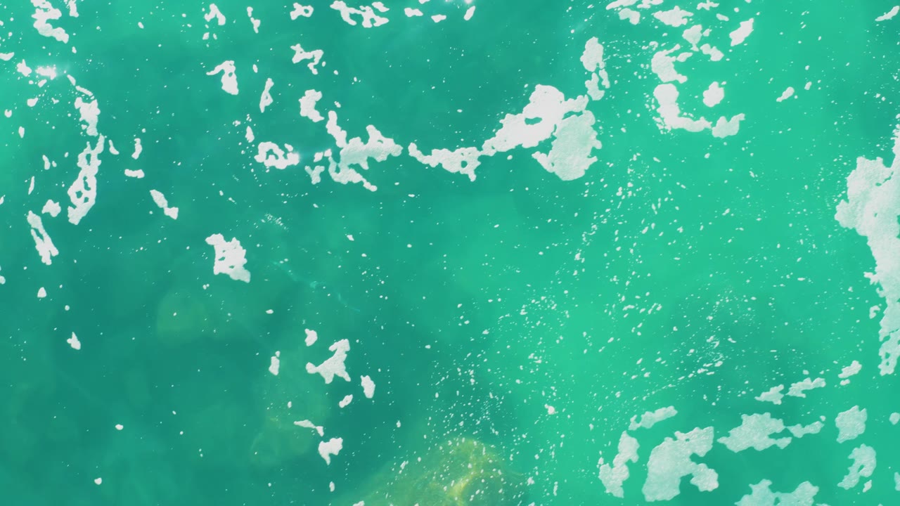 Aerial-style top-down frame at Laguna Beach Los Cabos with blue-green water, scattered white foam, a pale submerged patch near the lower area, and no visible people.