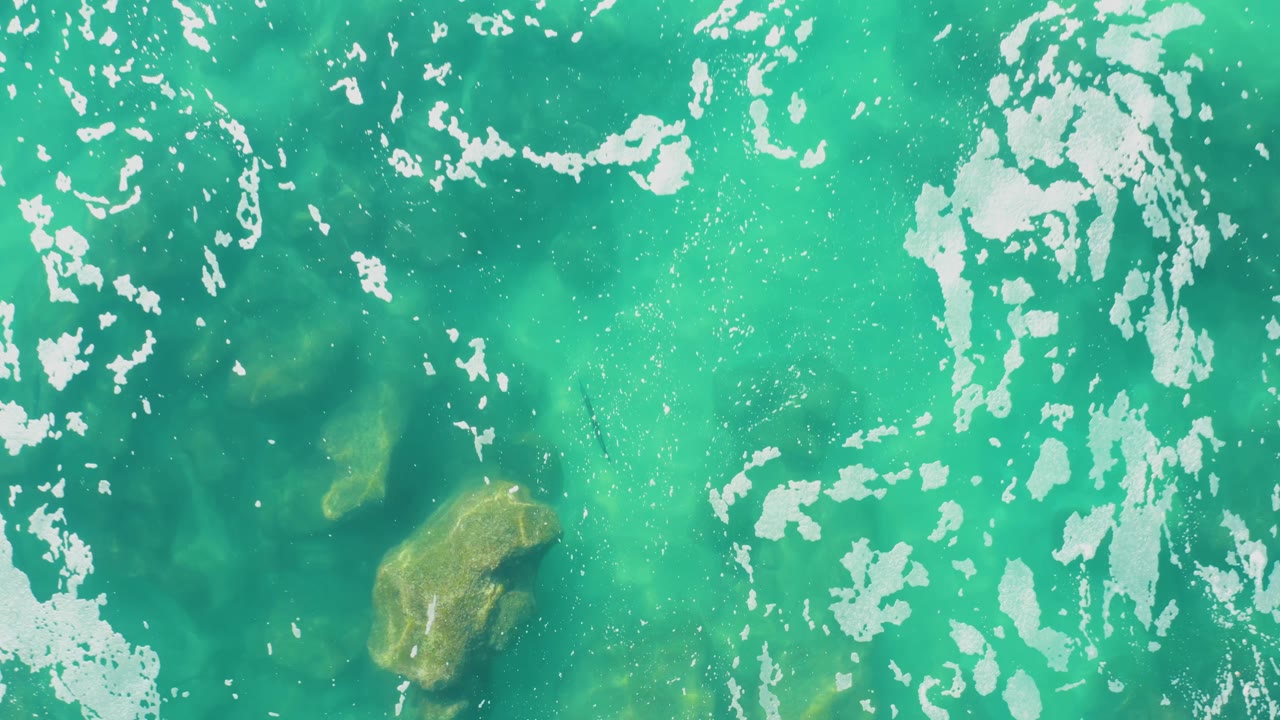 A second overhead frame shows turquoise water in Laguna Beach Los Cabos with foam clustered across the upper area, a larger submerged rock left of center, and no visible people.