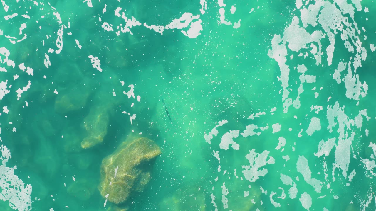Overhead turquoise ocean at Laguna Beach Los Cabos shows broken white foam across the top, darker water patches, a golden submerged rock near the lower right, and no visible people.