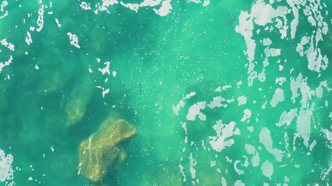 This overhead frame from Laguna Beach Los Cabos shows broad turquoise water, streaked white foam on the right, faint submerged rocks through the water, and no visible people.