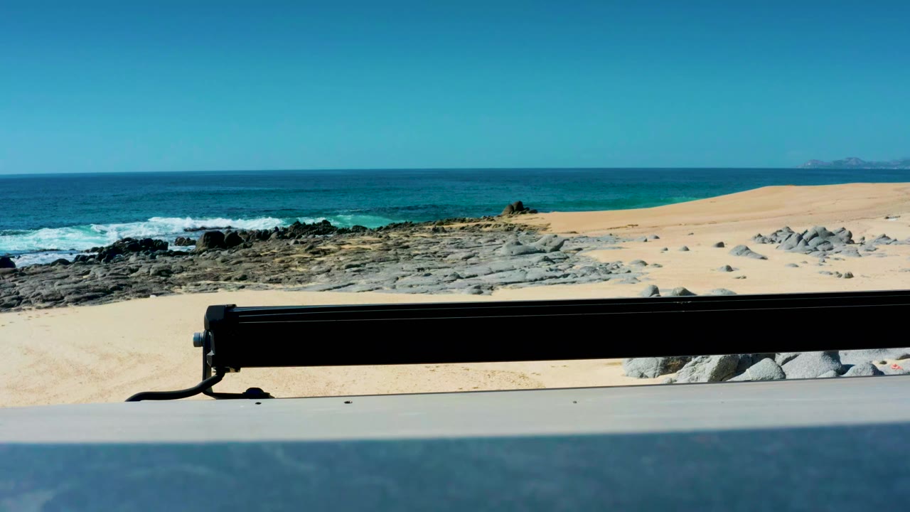 Laguna Beach Los Cabos beach scene with the black foreground rail still crossing the frame, waves breaking near dark rocks, and open sand under clear sky, no visible people.