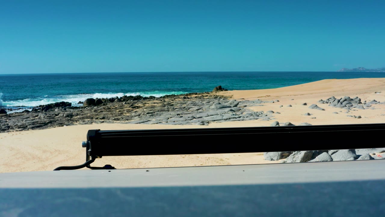 Ocean waves and rocky sand beach seen behind a black vehicle rail.