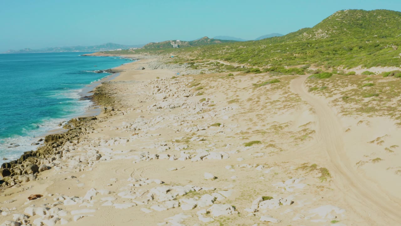 Elevated view of Laguna Beach Los Cabos with turquoise ocean at left, pale rocky sand across the beach, green hills at right, clear blue sky, and no visible people.