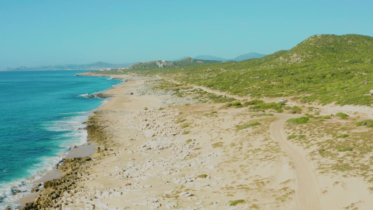 Laguna Beach Los Cabos coastline with turquoise water on the left, white surf, pale sand, rocky patches, a dirt track on the right, hillside greenery, and no visible people.
