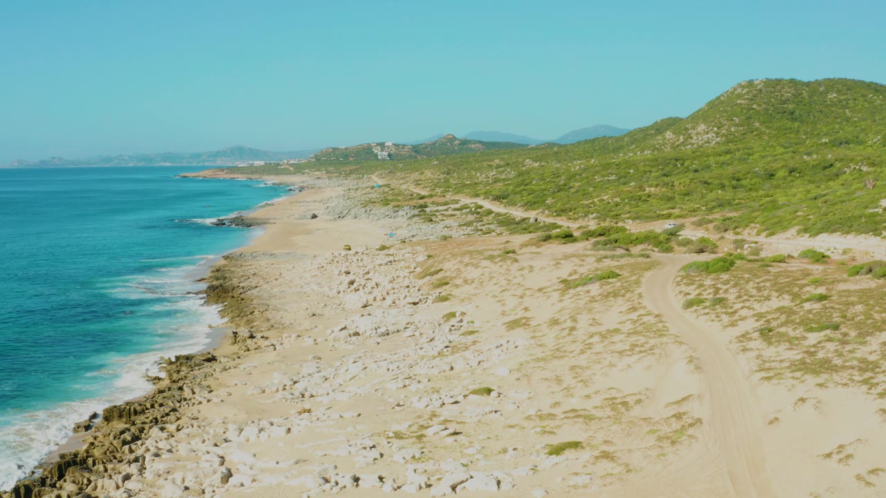 Aerial coastal frame from Laguna Beach Los Cabos showing turquoise ocean along the left edge, foamy surf, rocks on pale sand, green hillside to the right, and no visible people.