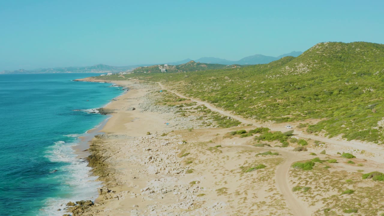 Laguna Beach Los Cabos shoreline with blue water meeting sand and rocks, a curving dirt path through the hillside, and no visible people.