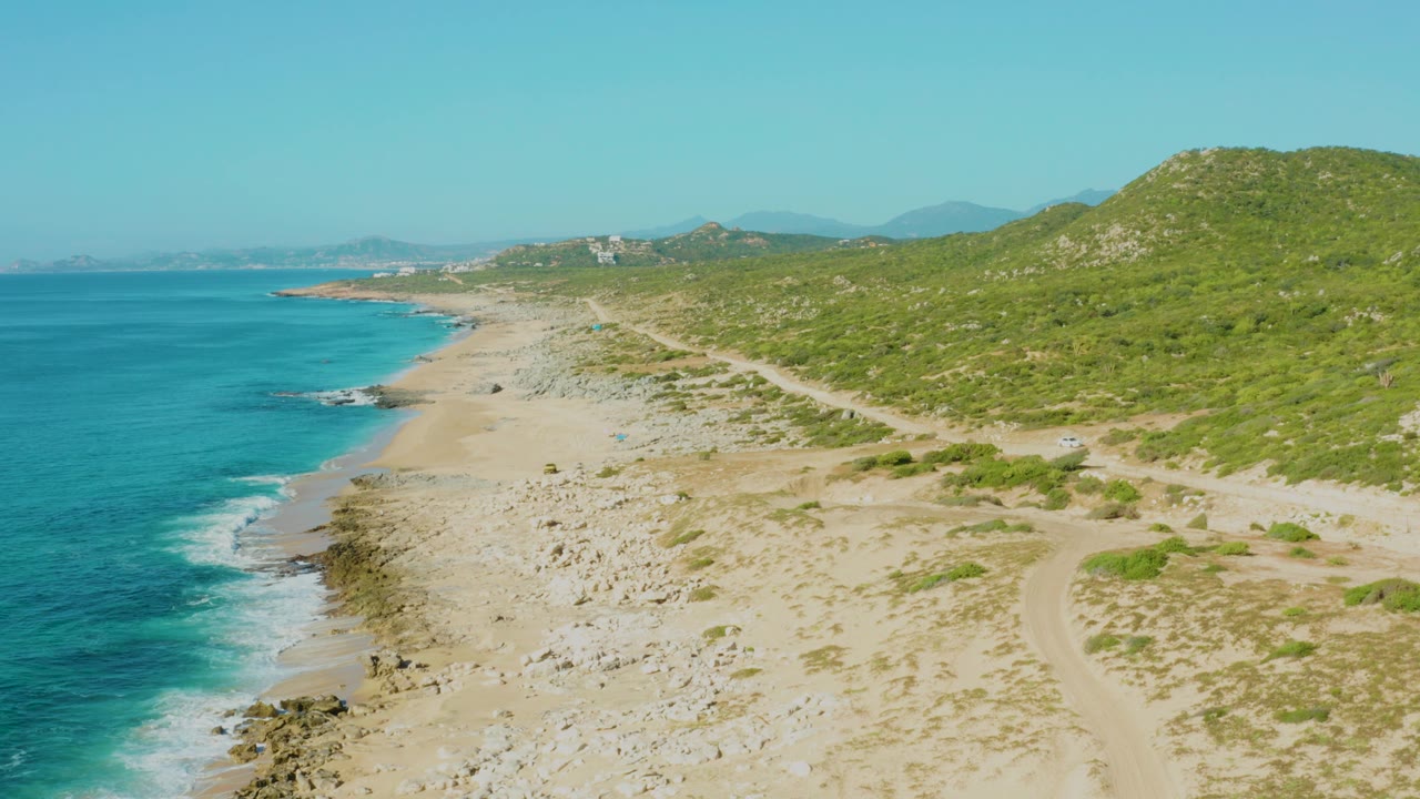 Mid-clip aerial view of Laguna Beach Los Cabos with waves breaking near rocks, a pale beach, hillside scrub, a dirt track, and no visible people.