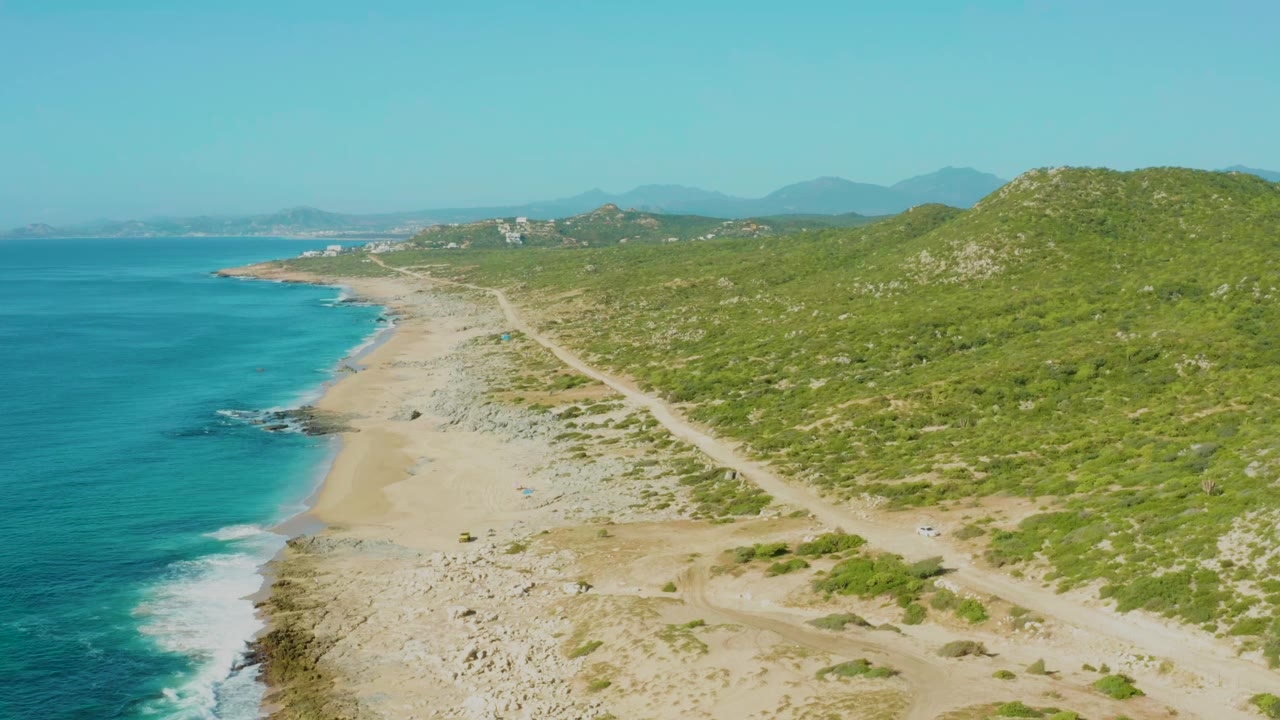 Aerial view at Laguna Beach Los Cabos with the waterline slightly farther down frame, foamy waves along the left shore, a dirt track through pale sand, green hillside scrub at right, and no visible people.